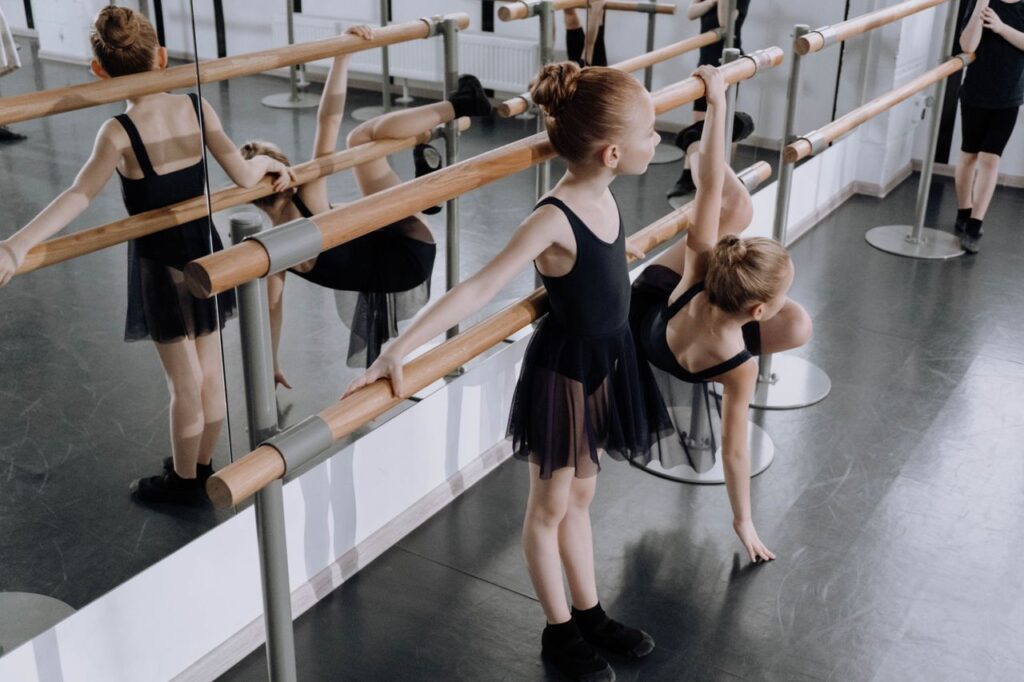 Two young ballerinas practice ballet poses at the barre in a studio, focusing on balance and flexibility.