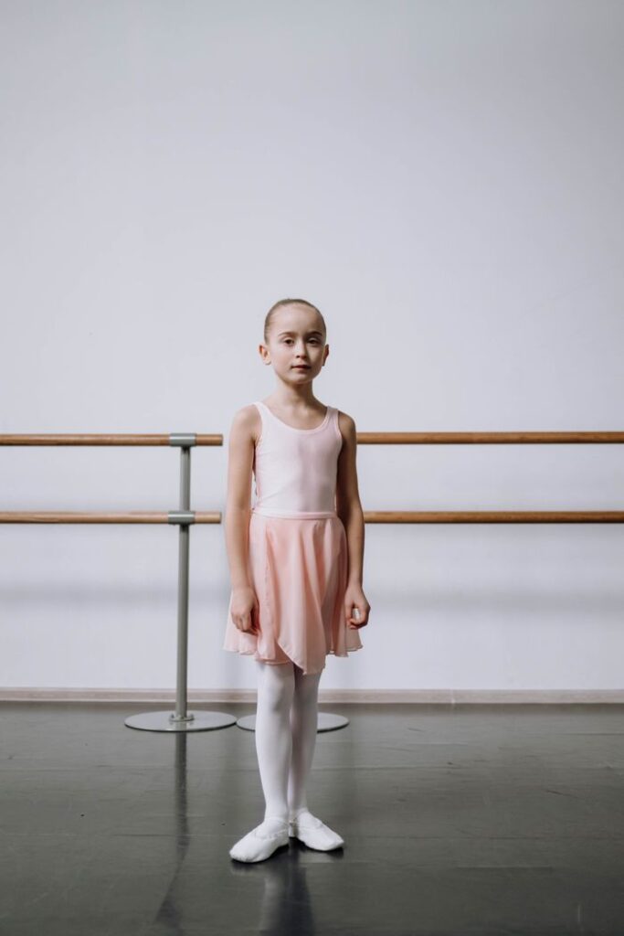 A young girl in ballet attire standing gracefully in a studio setting with a ballet barre.
